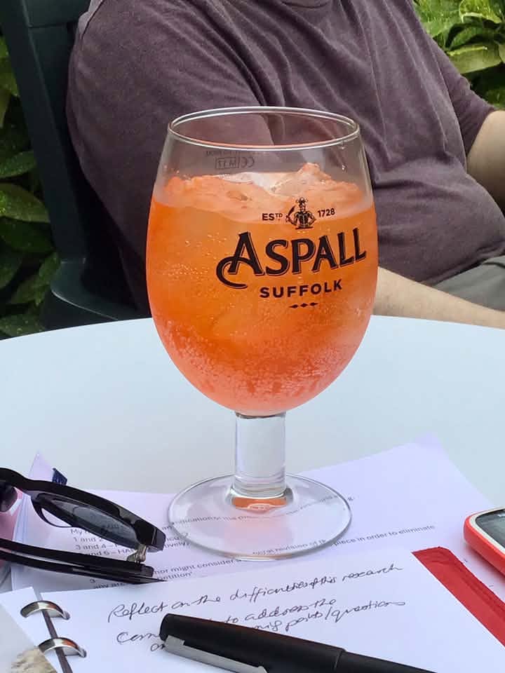 An orange coloured cocktail with ice served in a pint sized Aspall cider glass, with the torso of a person in the background
